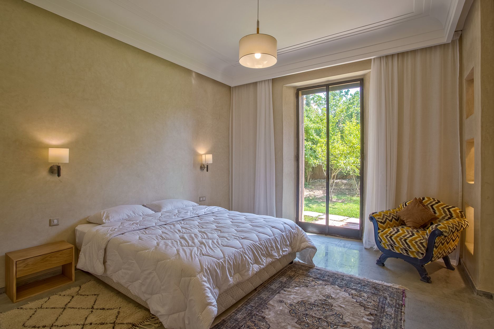 A bedroom with a large bed, a chair, and a view of greenery outside.