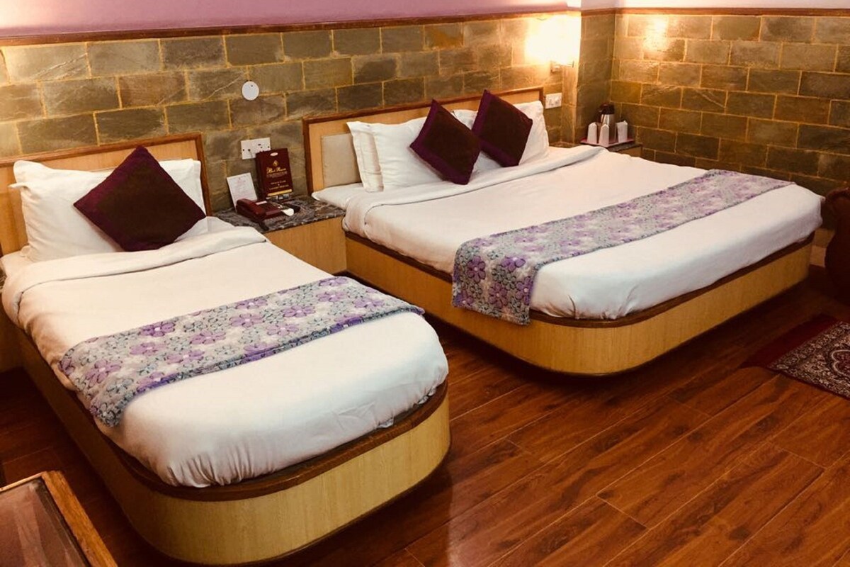 Dos camas individuales con cabeceros de madera, sábanas blancas y almohadas moradas. Se ve una mesita de noche con una lámpara y un libro. La habitación tiene una pared de piedra y suelo de madera.