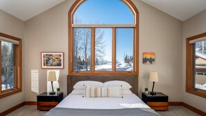 A bedroom with a large arched window, a bed with white bedding, two bedside tables with lamps, and framed artwork on the walls.