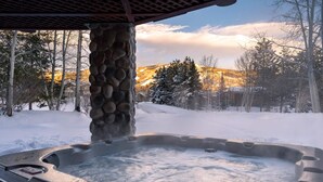 A hot tub with a stone pillar and a view of snow-covered trees and mountains.