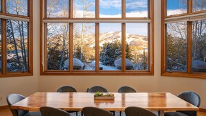A conference room with a large wooden table and six chairs, large windows with a view of snow-covered mountains and trees.