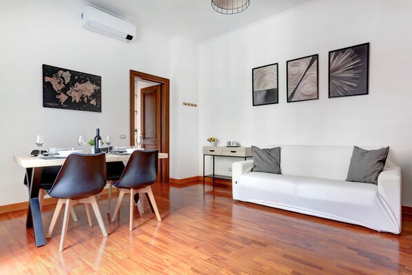 A modern living room with a white sofa, two black chairs, a wooden dining table with chairs, a small desk, and a world map on the wall. - Colosseo Green Park – Home and More (Rome)