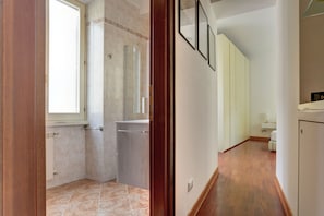 A hallway with a bathroom on the left, featuring a shower, a toilet, and a sink. The hallway has wooden flooring and white walls. - Colosseo Green Park – Home and More (Rome)