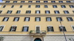 Un immeuble à plusieurs étages, doté d’une façade jaune, de nombreuses fenêtres avec volets et d’une entrée centrale surmontée d’une arche décorative.