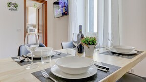 Une table à manger est dressée avec de l’assiette blanche, des bols, des verres à vin et des couverts. Une bouteille de vin et une petite plante en pot sont posées sur la table.