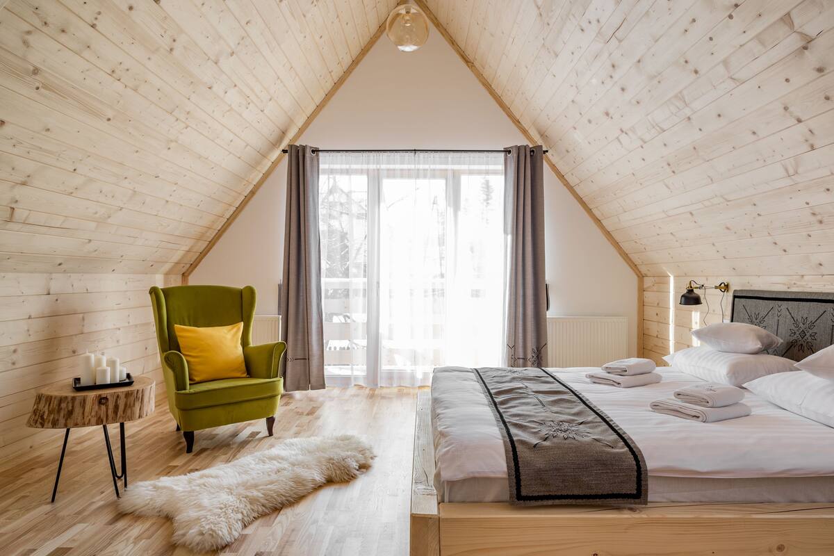 A cozy bedroom with a sloped ceiling, a wooden bed, a green armchair, a fluffy rug, and a small side table with candles.