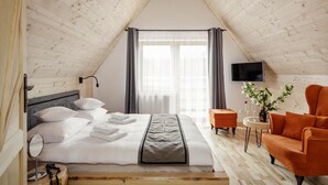 A bedroom with a sloped ceiling, a bed with white linens, a grey throw, a small orange armchair, a side table with a bottle and glasses, and a TV on the wall. - Tatra Wood House (Zakopane)