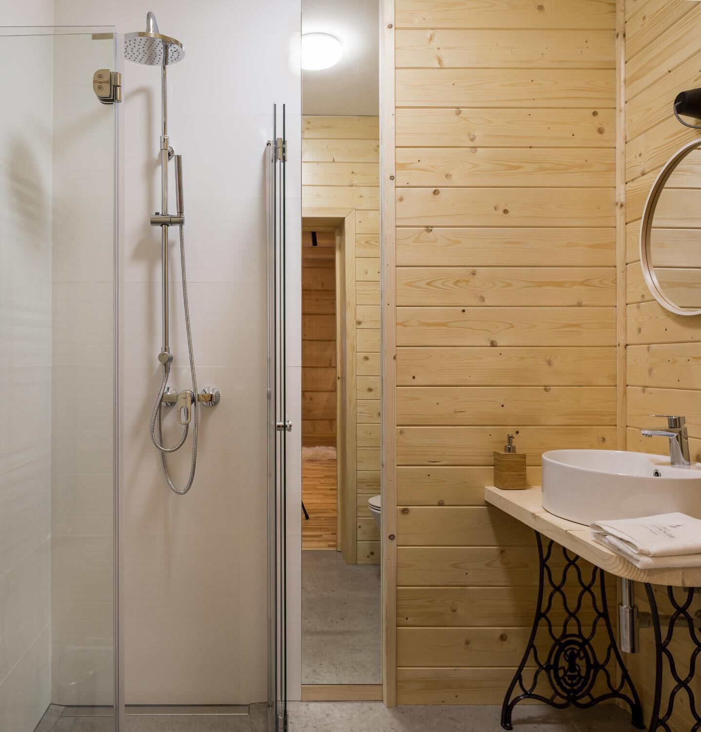 Ett modernt badrum med en dusch som är avskärmad med glasväggar, en väggpanel i trä, en rund spegel och en vit tvättställ med en svart metallram.