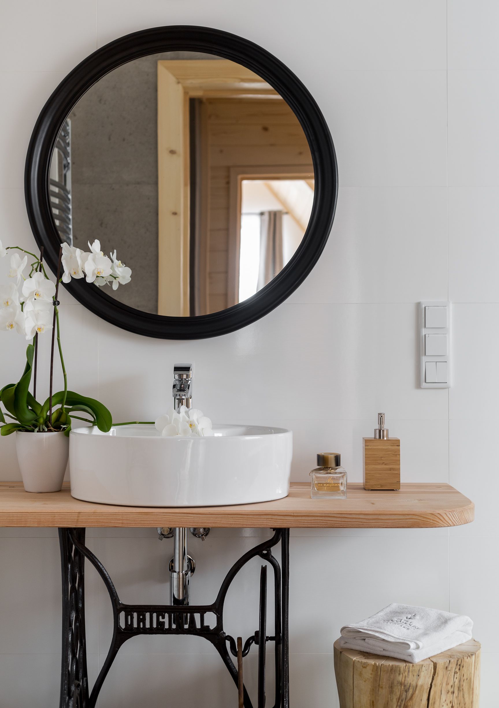 A bathroom with a white sink, a black-framed mirror, a potted orchid, a wooden soap holder, and a towel on a wooden stool.