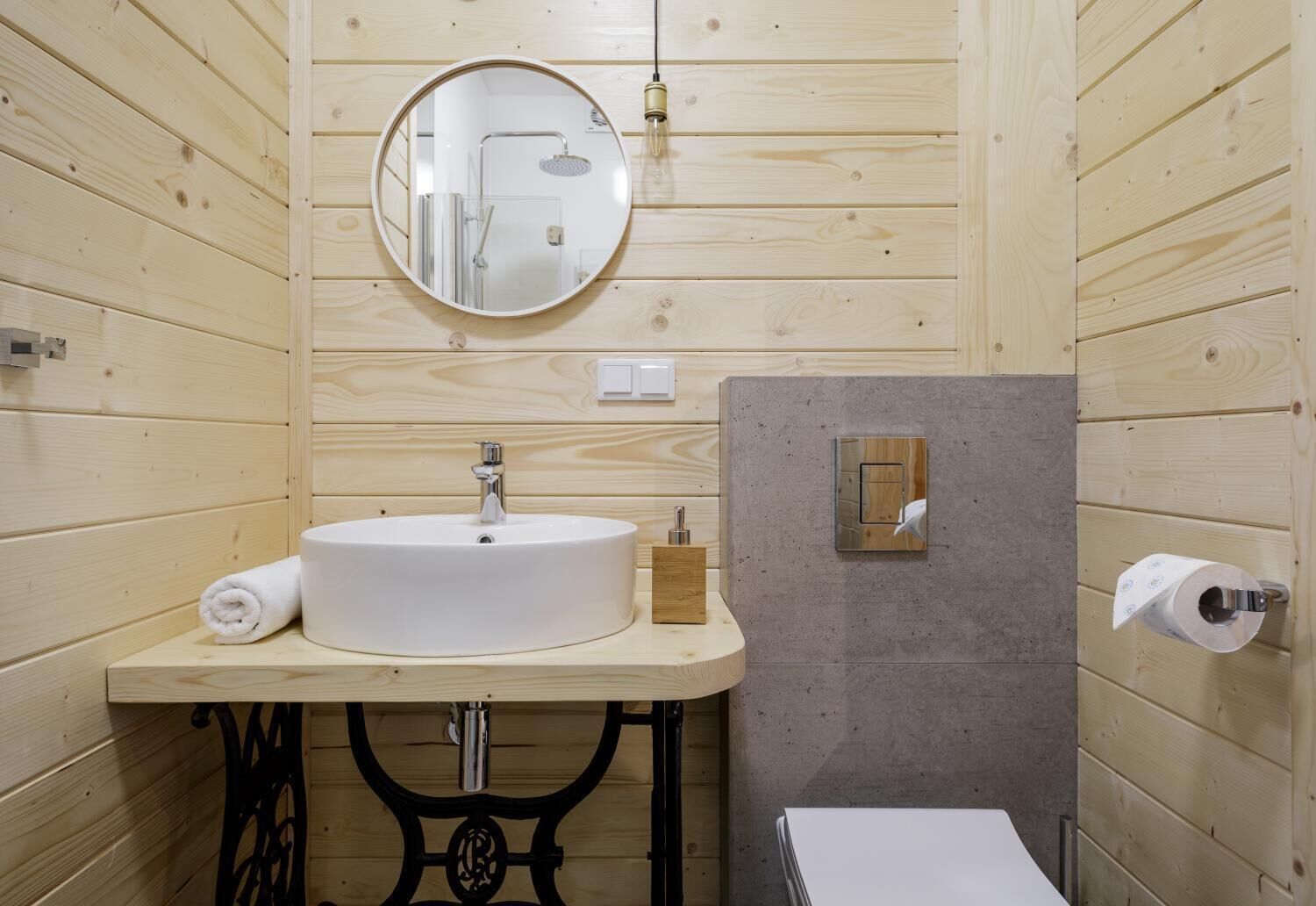 A wooden-paneled bathroom with a round mirror, a sink, a toilet, and a towel.