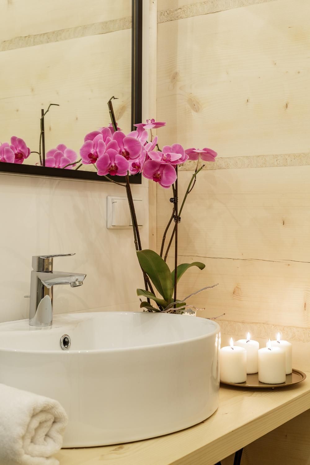A bathroom with a round white sink, a mirror, pink orchids, and white candles on a wooden shelf.