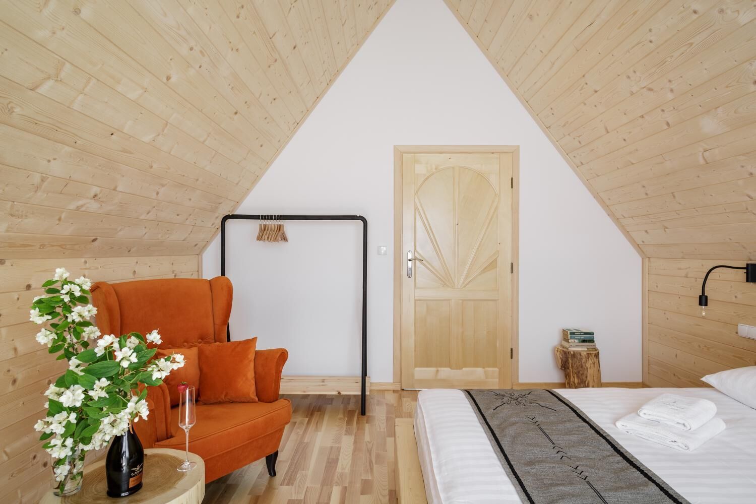 Ein gemütliches Schlafzimmer unter dem Dach mit Holzbalkendecke, einer Holztür, einem Bett mit weißen Bettwäsche, einem kleinen Nachttisch mit Flasche und Glas sowie einem orangefarbenen Sessel.