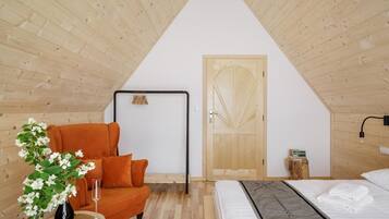 Ein gemütliches Schlafzimmer unter dem Dach mit Holzbalkendecke, einer Holztür, einem Bett mit weißen Bettwäsche, einem kleinen Nachttisch mit Flasche und Glas sowie einem orangefarbenen Sessel.