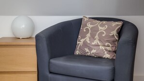 A dark grey armchair with a patterned cushion, a wooden nightstand with a white spherical object, and a grey carpet. - The Annexe (Cheltenham)