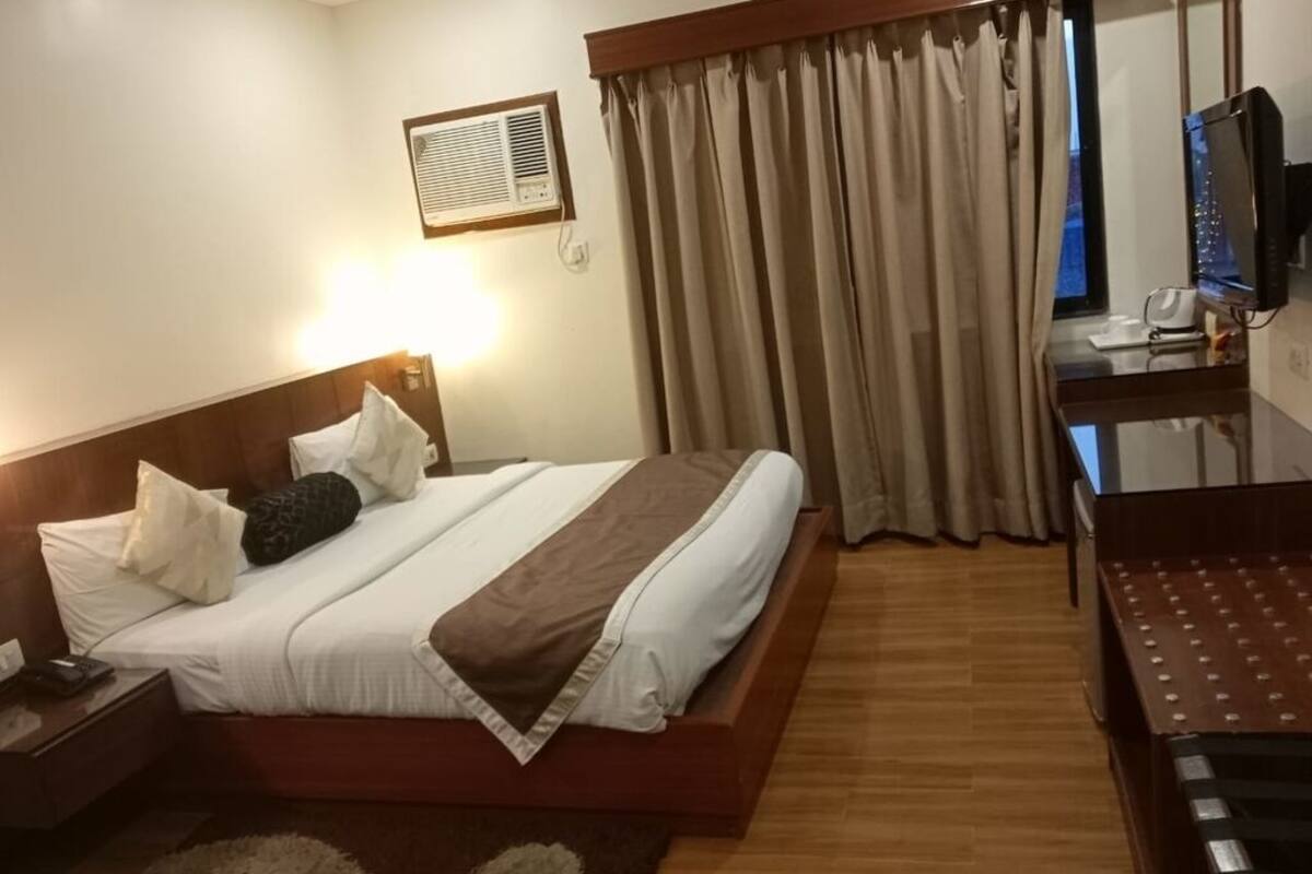 Habitación de hotel con cama, escritorio con computadora, ventana con cortinas y unidad de aire acondicionado de pared.
