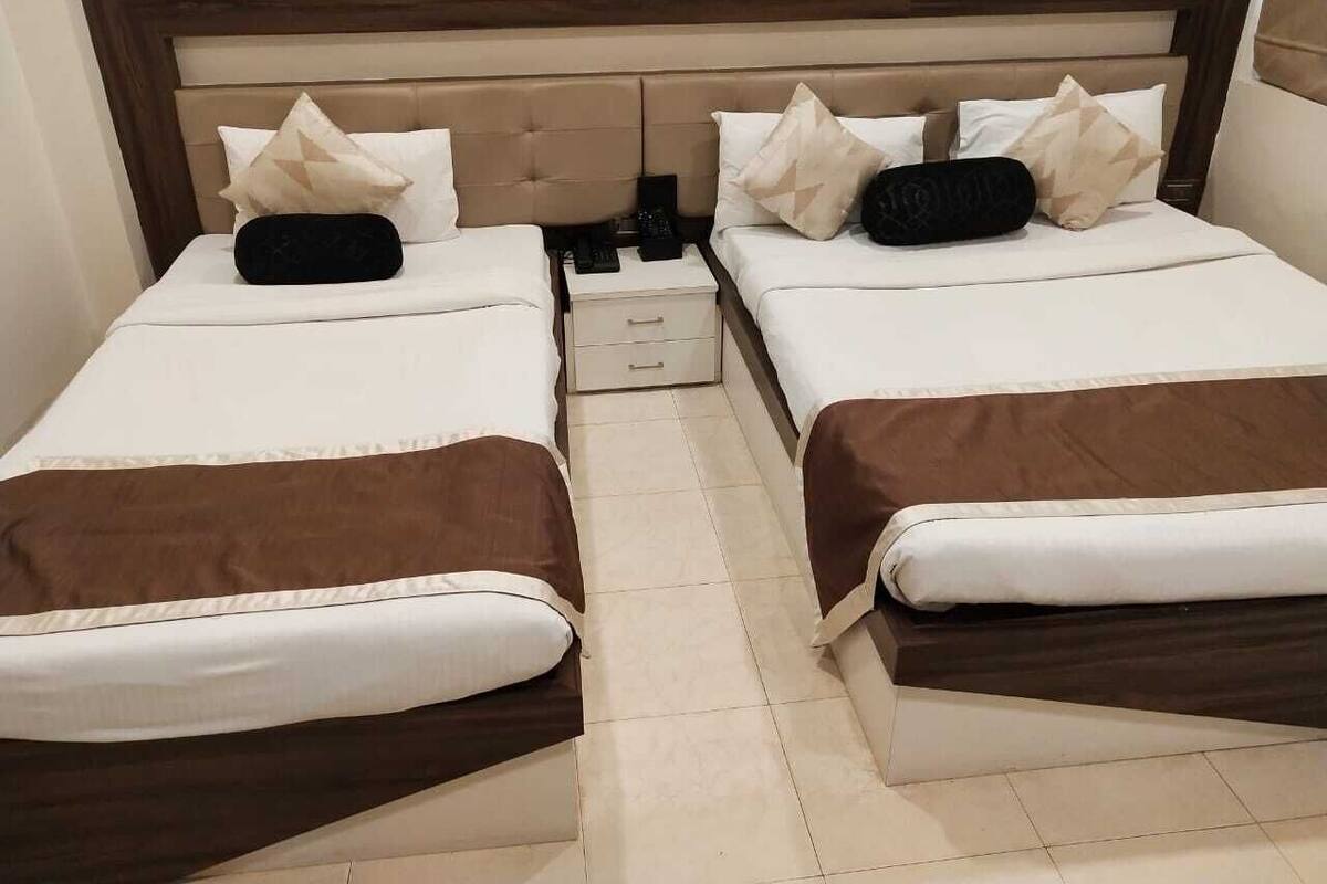 Habitación doble con dos camas, cada una con cabecero marrón, ropa de cama blanca y almohadas decorativas. Un pequeño mesita de noche con una lámpara y un control remoto.