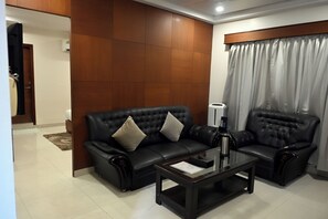 A modern living room with a black leather sofa, a glass coffee table, and a large window with curtains. - Hotel Baidyanath (Deoghar)