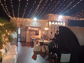 Un espacio de comedor al aire libre con luces colgantes, un letrero que dice 'Prangan' y mesas dispuestas para comer.