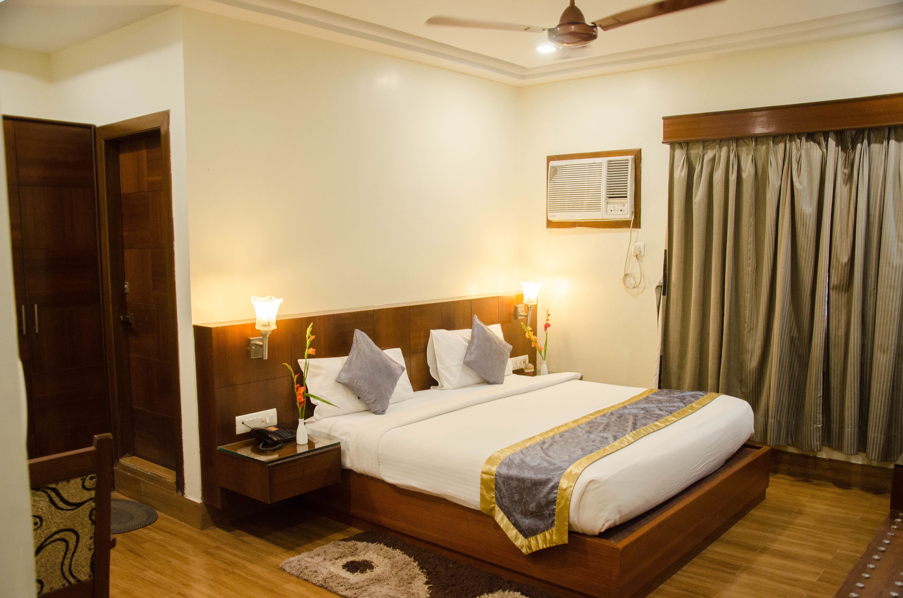 Habitación de hotel ordenada con una cama grande, lámparas de noche, ventilador de techo y una ventana con cortinas.