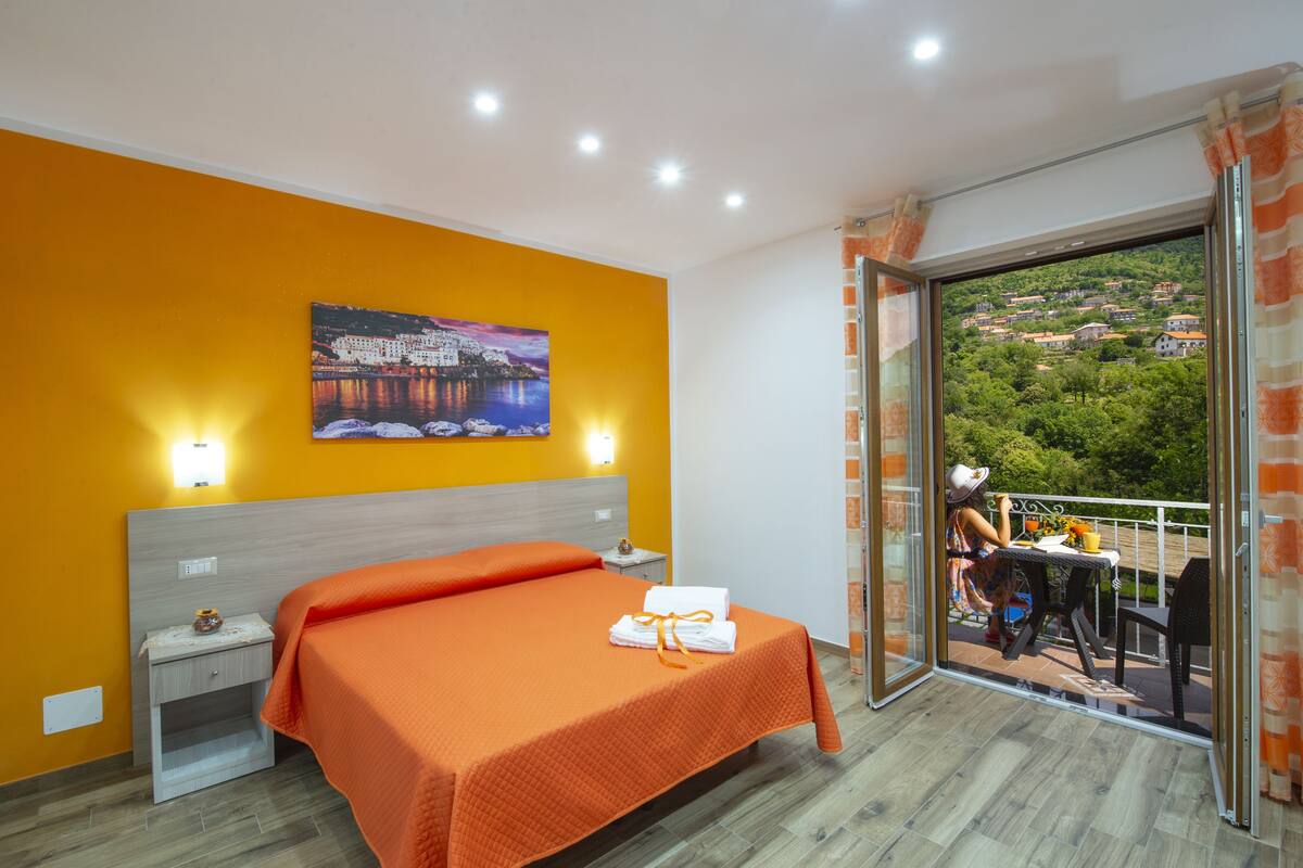 Une chambre d’hôtel avec une couvre-lit orange, un tableau représentant un paysage urbain, un balcon donnant sur un paysage verdoyant et une table avec une chaise.