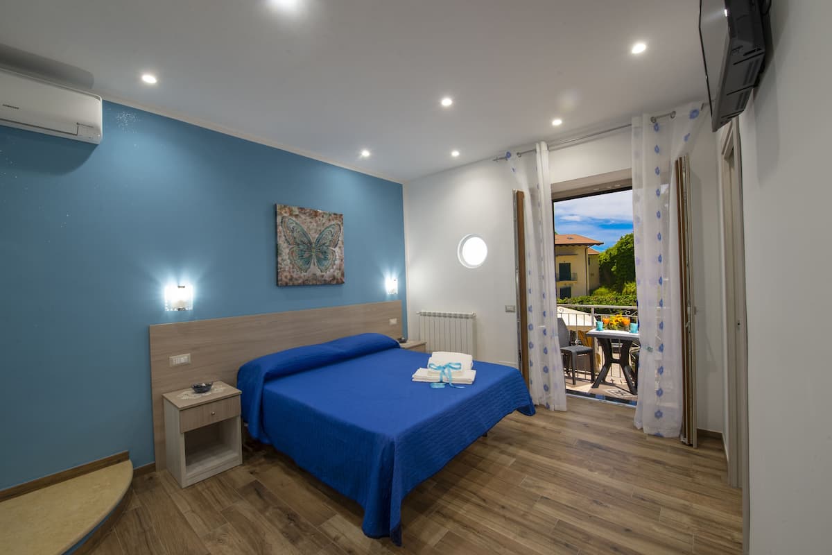 Une chambre avec une couvre-lit bleue, une table de chevet, un climatiseur fixé au mur, un luminaire au plafond et une vue sur l’extérieur par une porte.