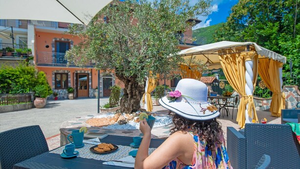An outdoor seating area with tables and chairs, a tree, and a building in the background. - B&B Il Girasole (Agerola)