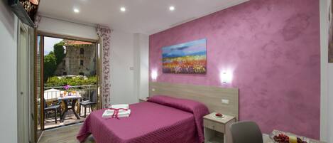 Un dormitorio con cama, mesita de noche, silla, mesa con frutero, un cuadro en la pared y balcón con mesa y sillas.