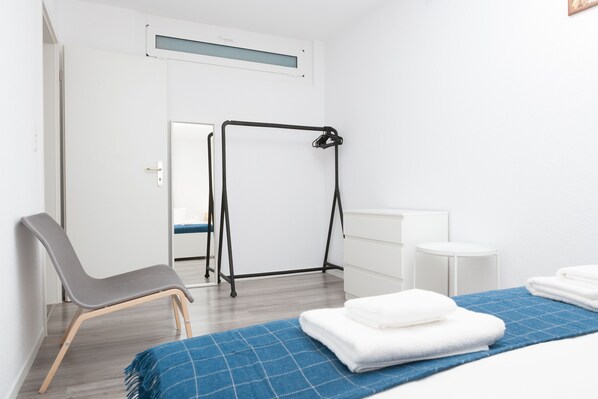 A hotel room with a bed, a chair, a small table, a wall-mounted rack, and a door leading to another room.