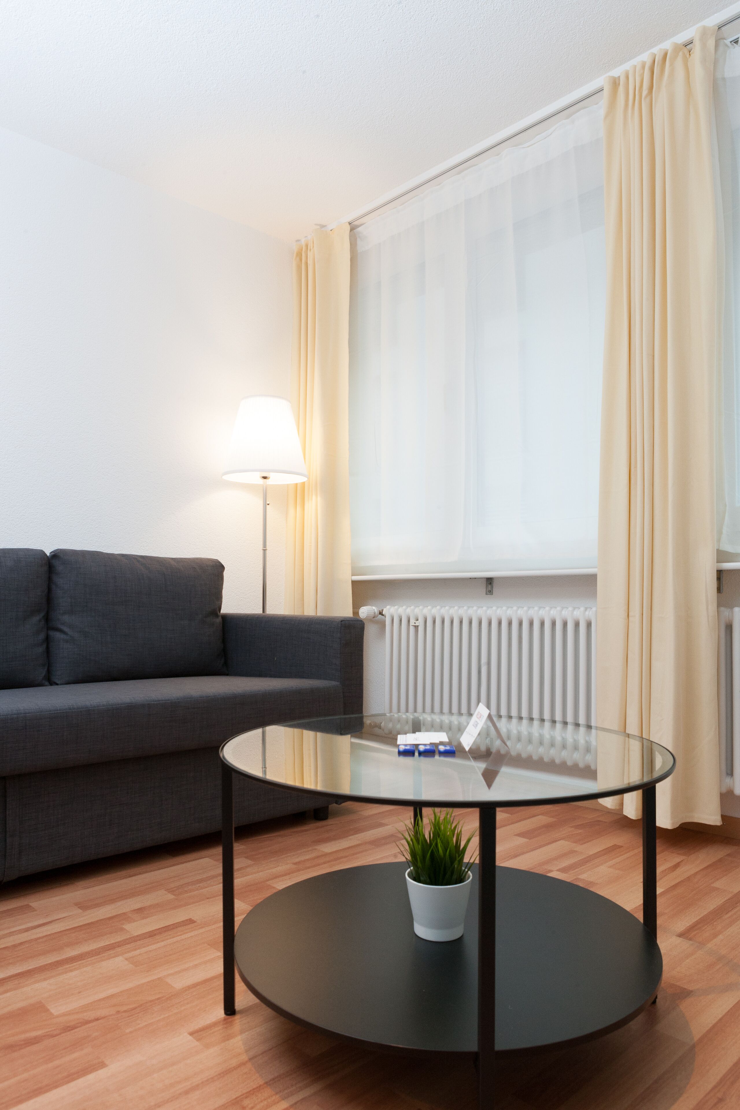 Ein modernes Wohnzimmer mit einem runden Glastisch, einem grauen Sofa, einer Stehlampe und Blick durch transparente Vorhänge.