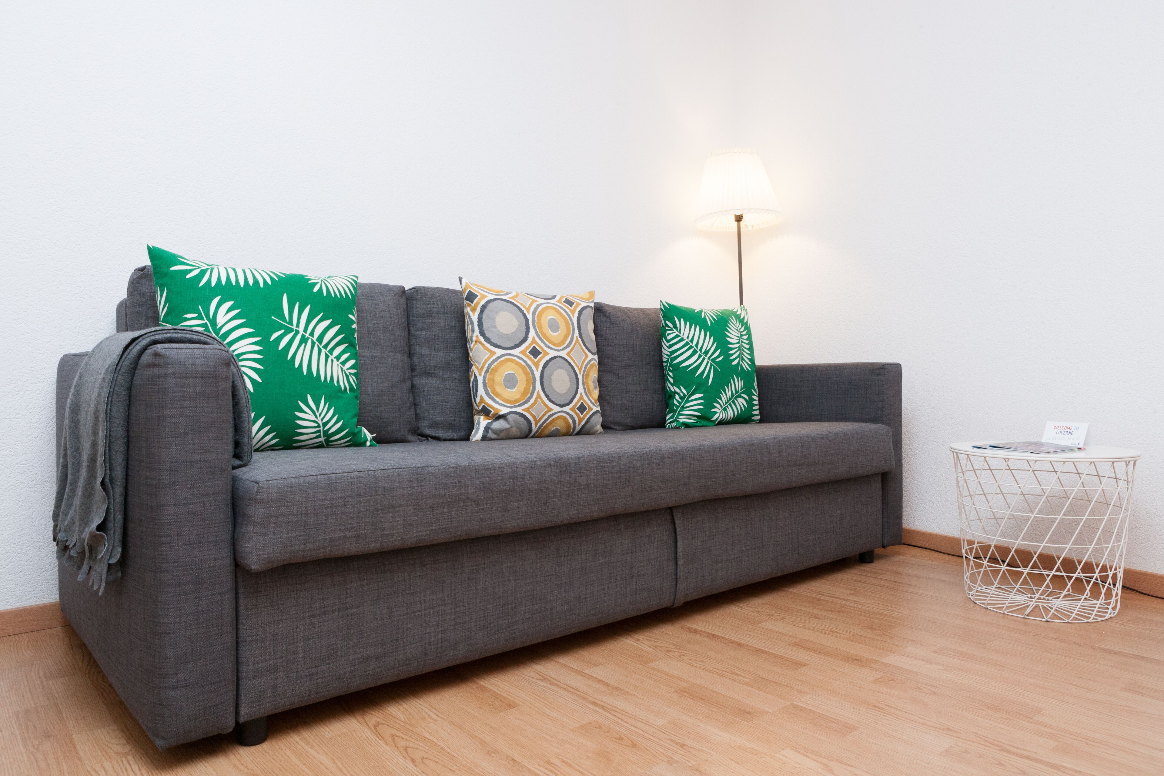 A grey sofa with patterned cushions, a lamp, and a wire basket on a wooden floor.