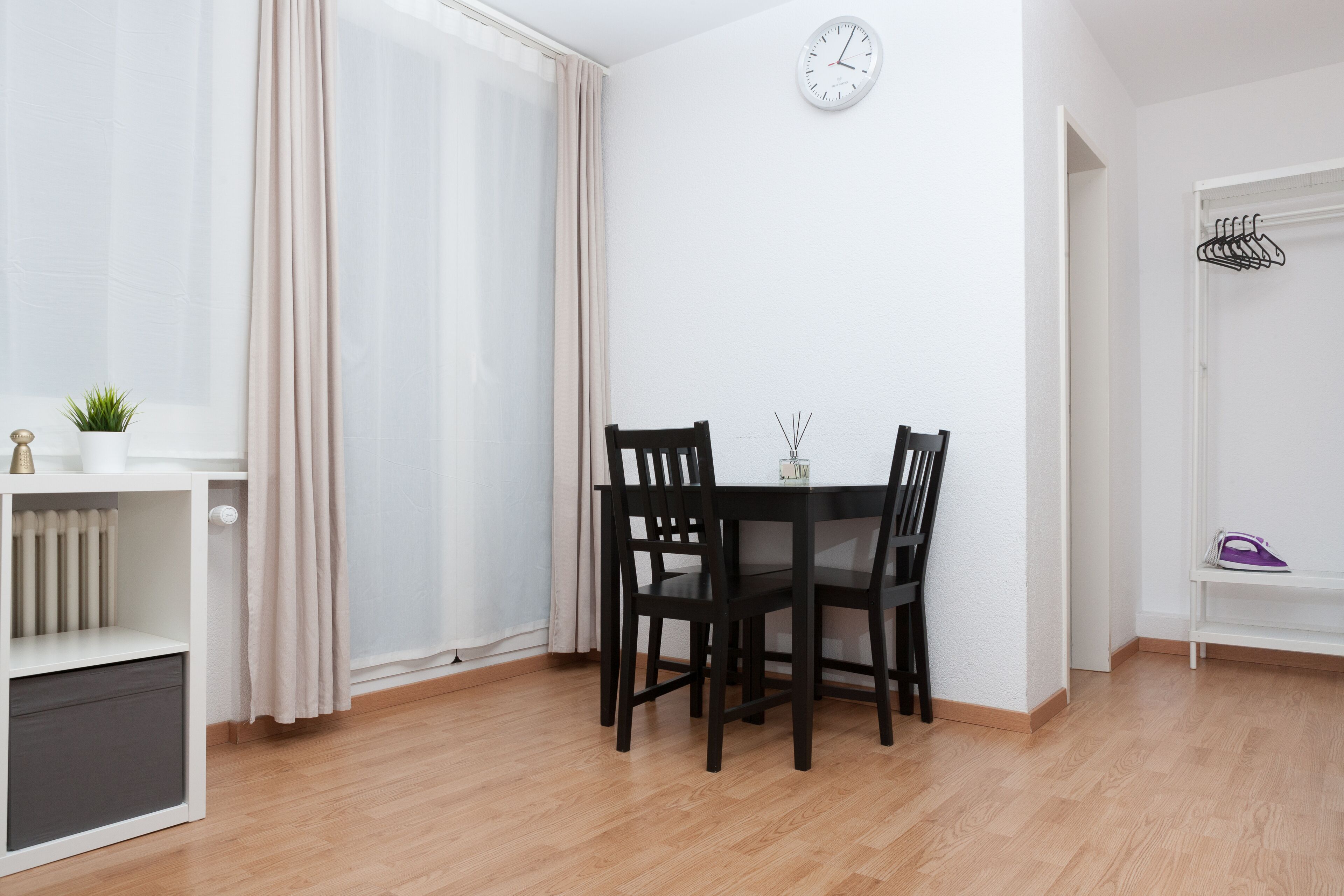 A room with a dining table and chairs, a clock on the wall, a radiator, a plant, and a coat rack.