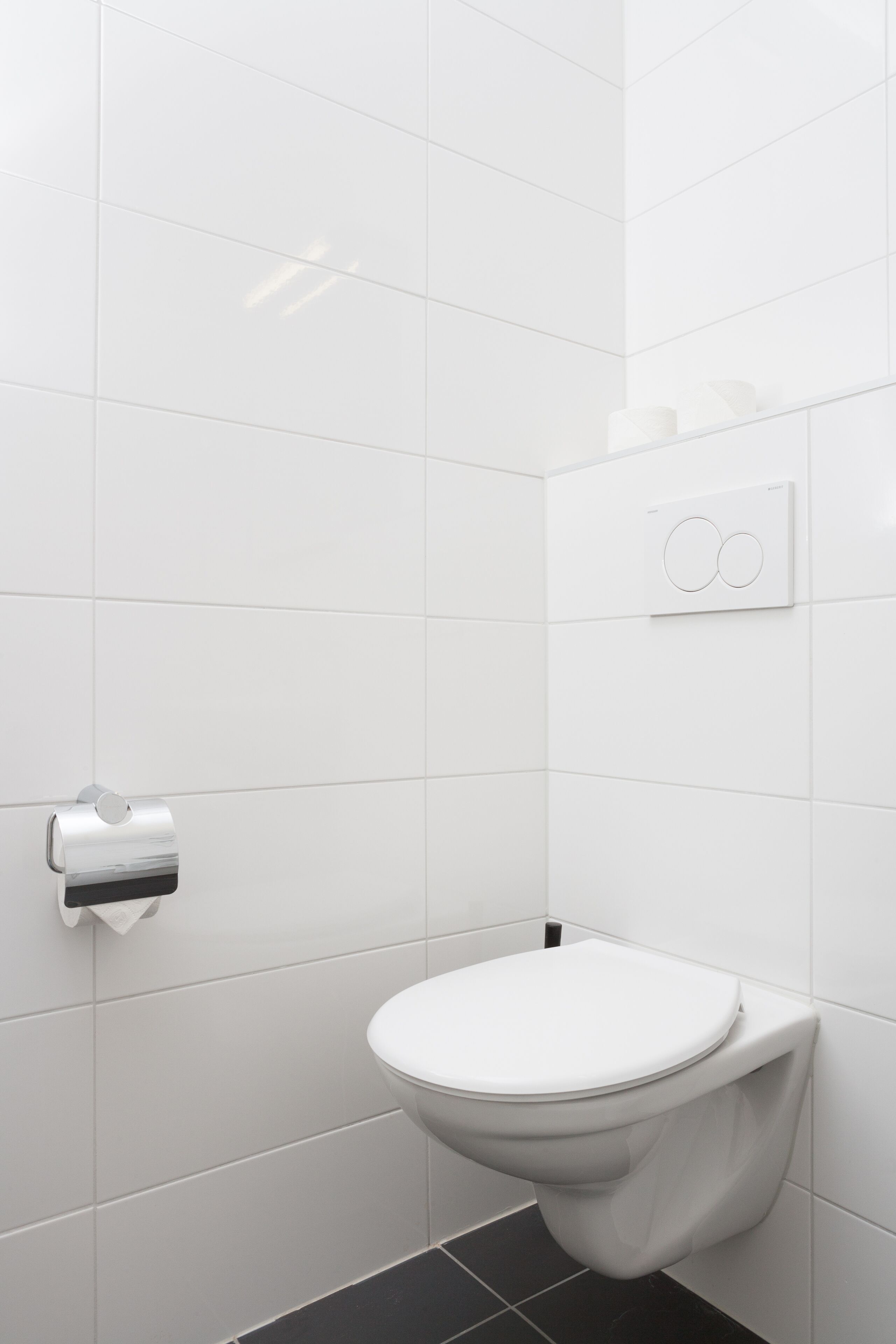Un baño moderno con azulejos blancos, un inodoro, un dispensador de toallas de papel y un botón para la descarga.