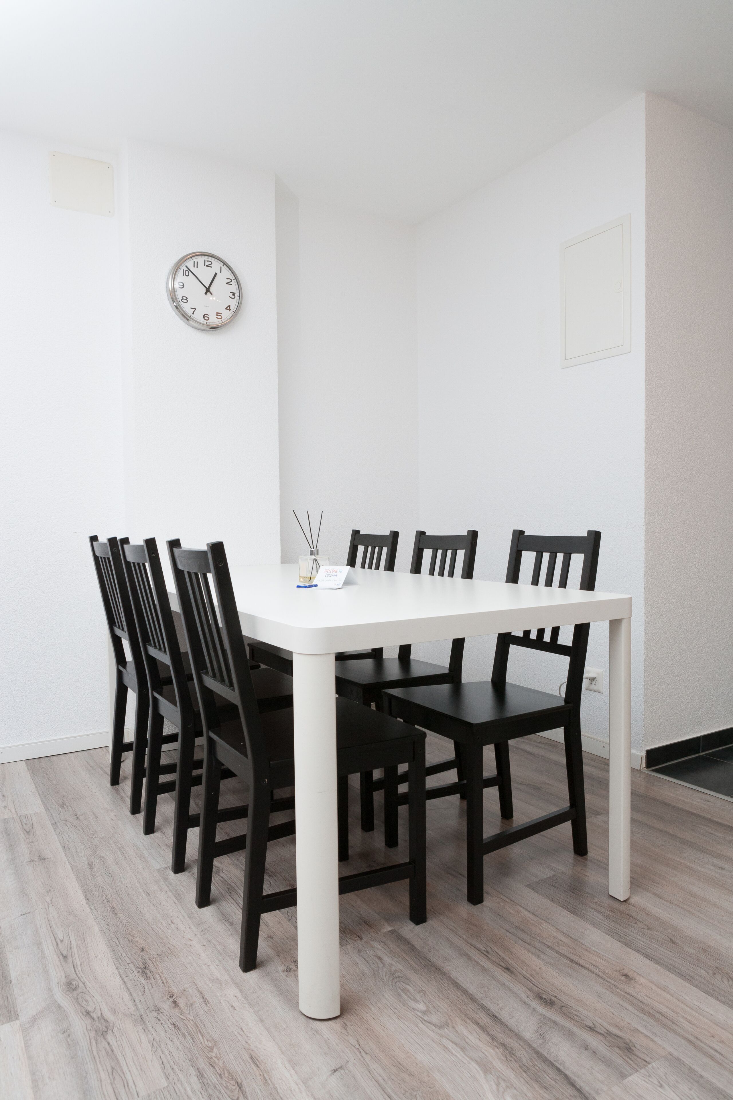 A dining area with a white table and black chairs, a clock on the wall, and a small vase with candles.