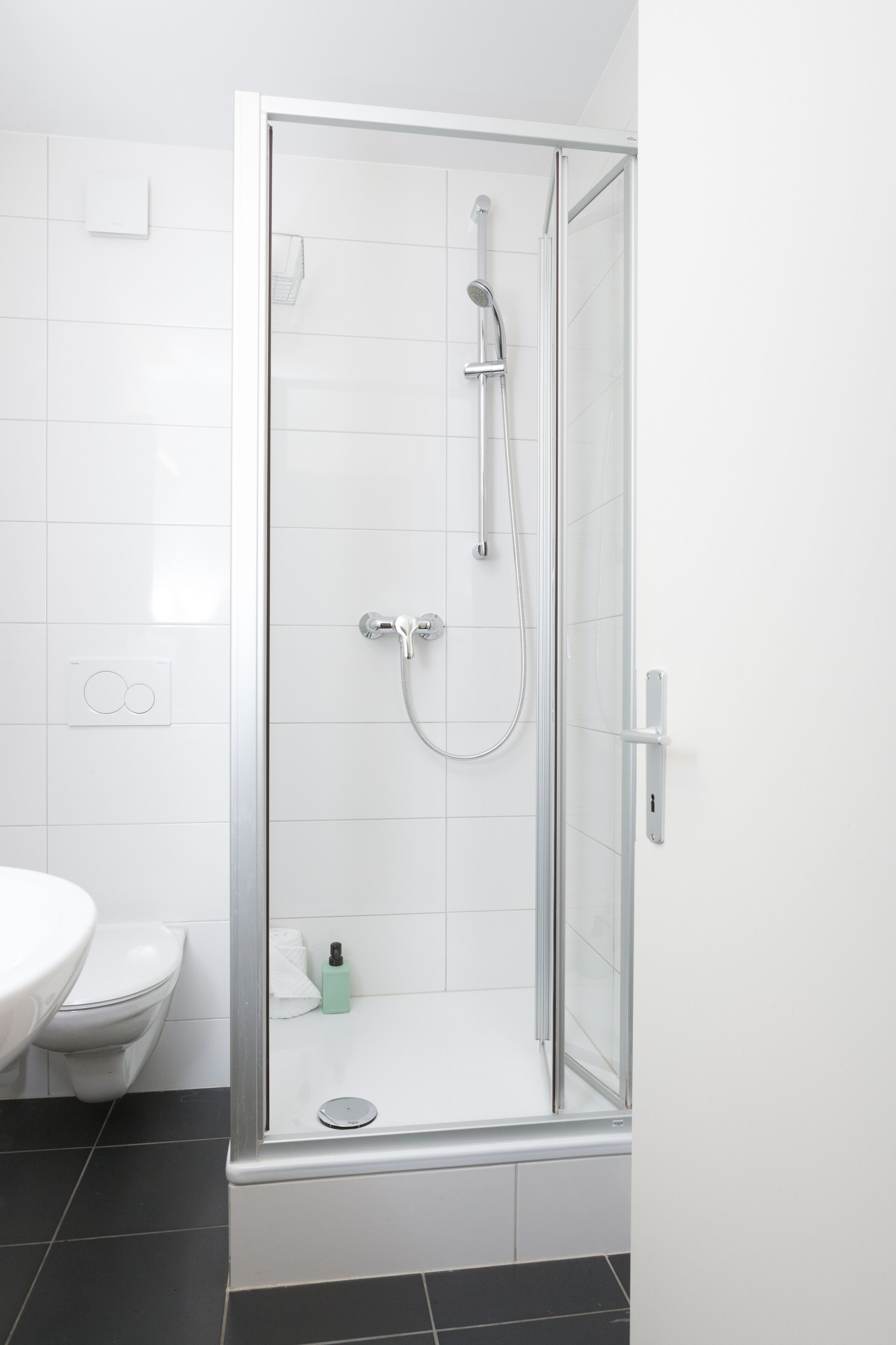 Et moderne badeværelse med en brusekabine i glas, hvide fliser, en hvid vask og et sort gulv.