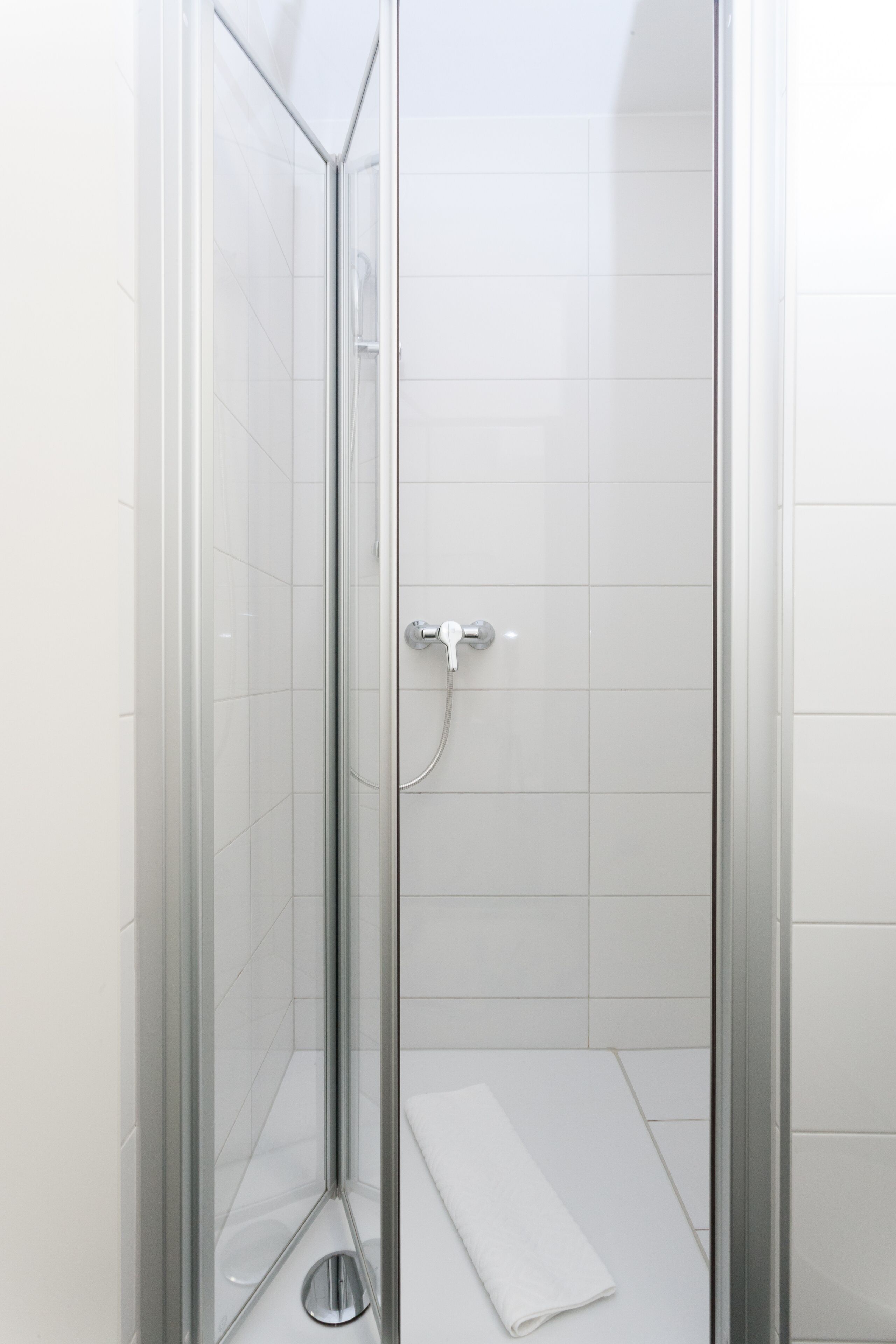 A glass-enclosed shower with a white tiled floor and a white towel on the floor.