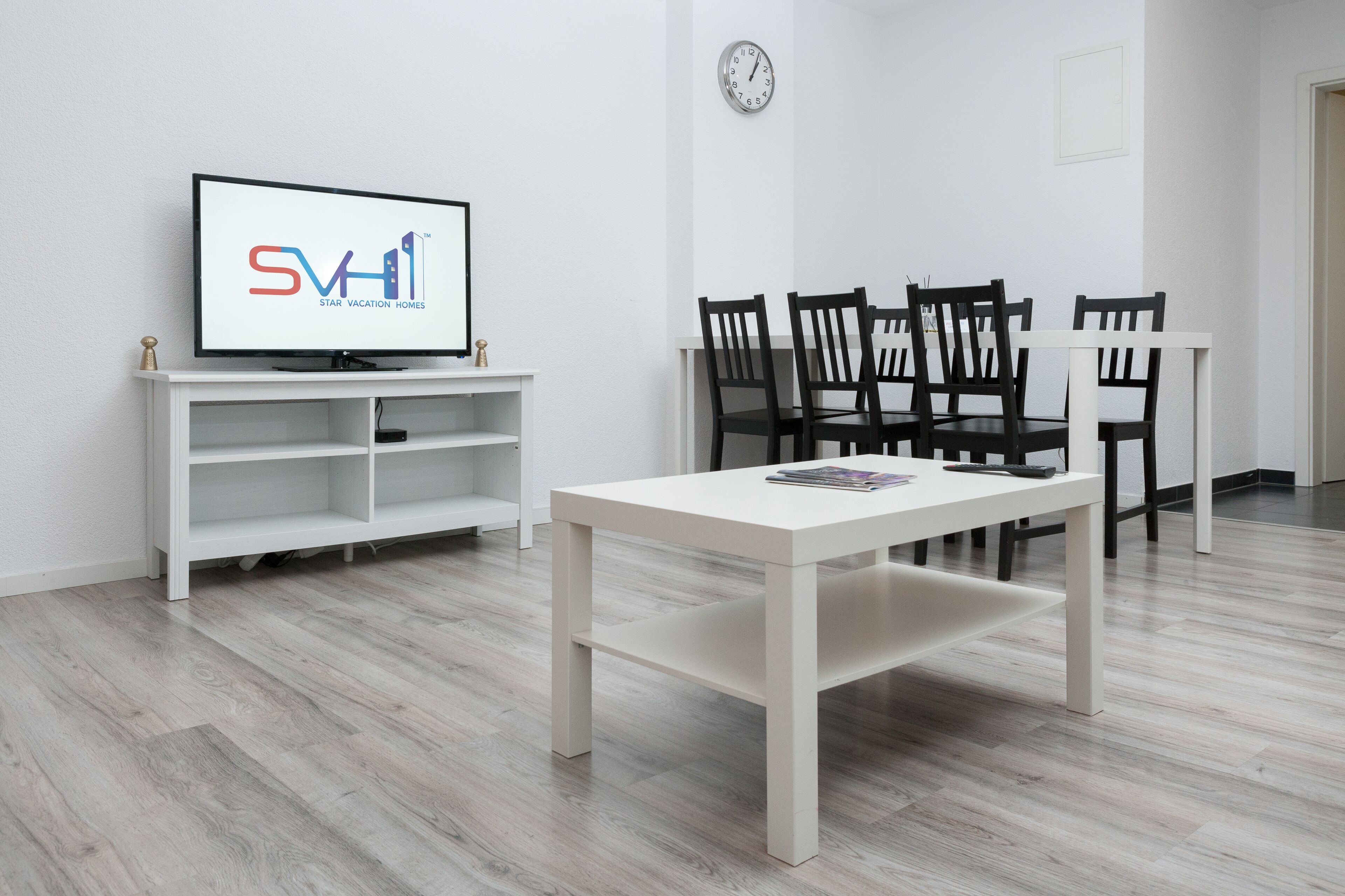 Ein modernes Zimmer mit Fernseher, der 'SVH Star Vacation Homes' anzeigt, einem weißen Tisch und schwarzen Stühlen.