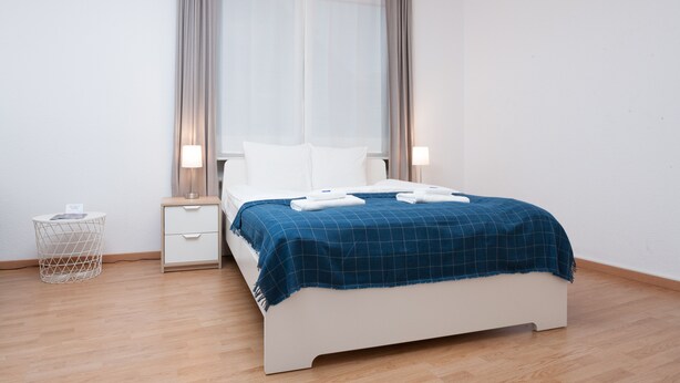 A modern hotel room with a large bed, a bedside table, a small side table, and a window with curtains. - Galaxy Apartments (Lucerne)