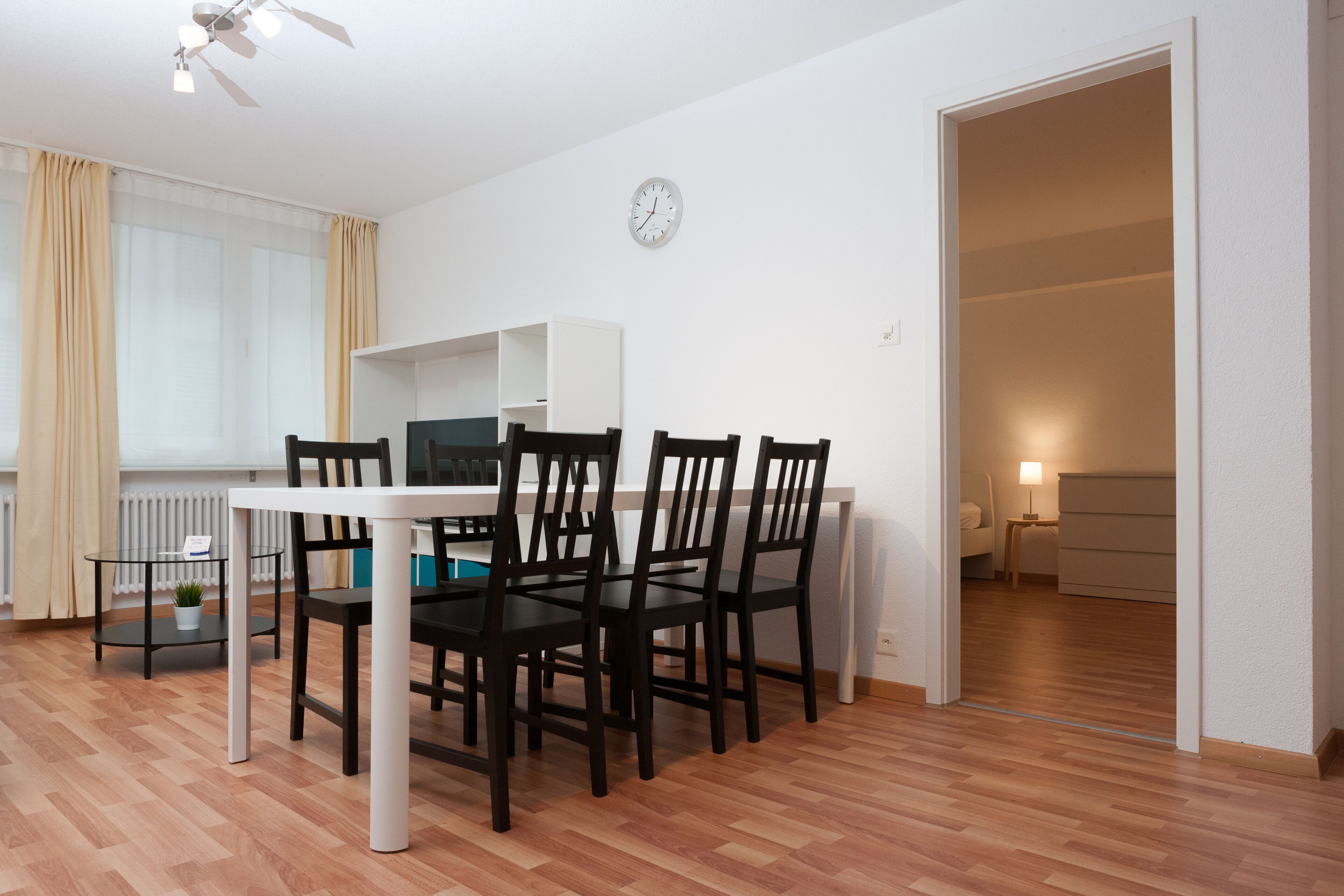 Uma área de refeições com mesa e cadeiras, um relógio na parede e vista para outro cômodo.