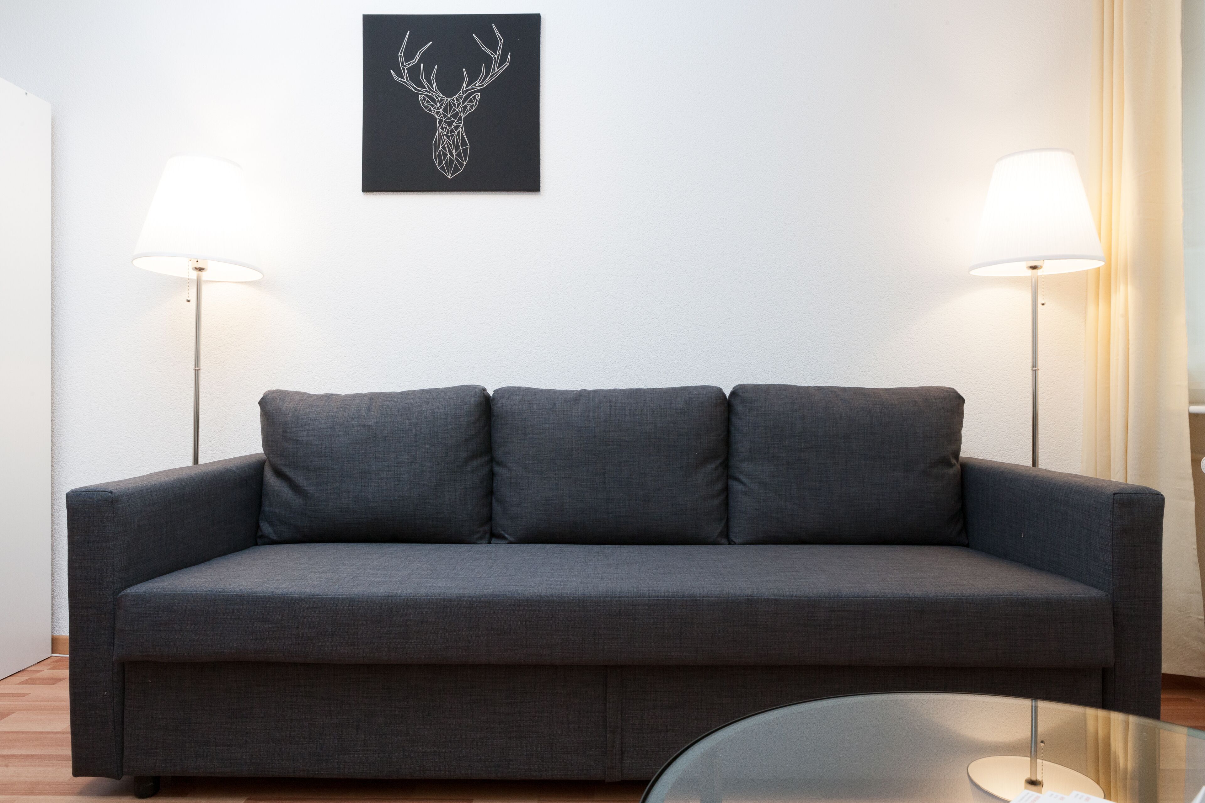 Ein graues Sofa mit zwei Lampen an beiden Seiten, einem Kunstwerk mit schwarzem Rahmen darüber und einem Glastisch im Vordergrund.