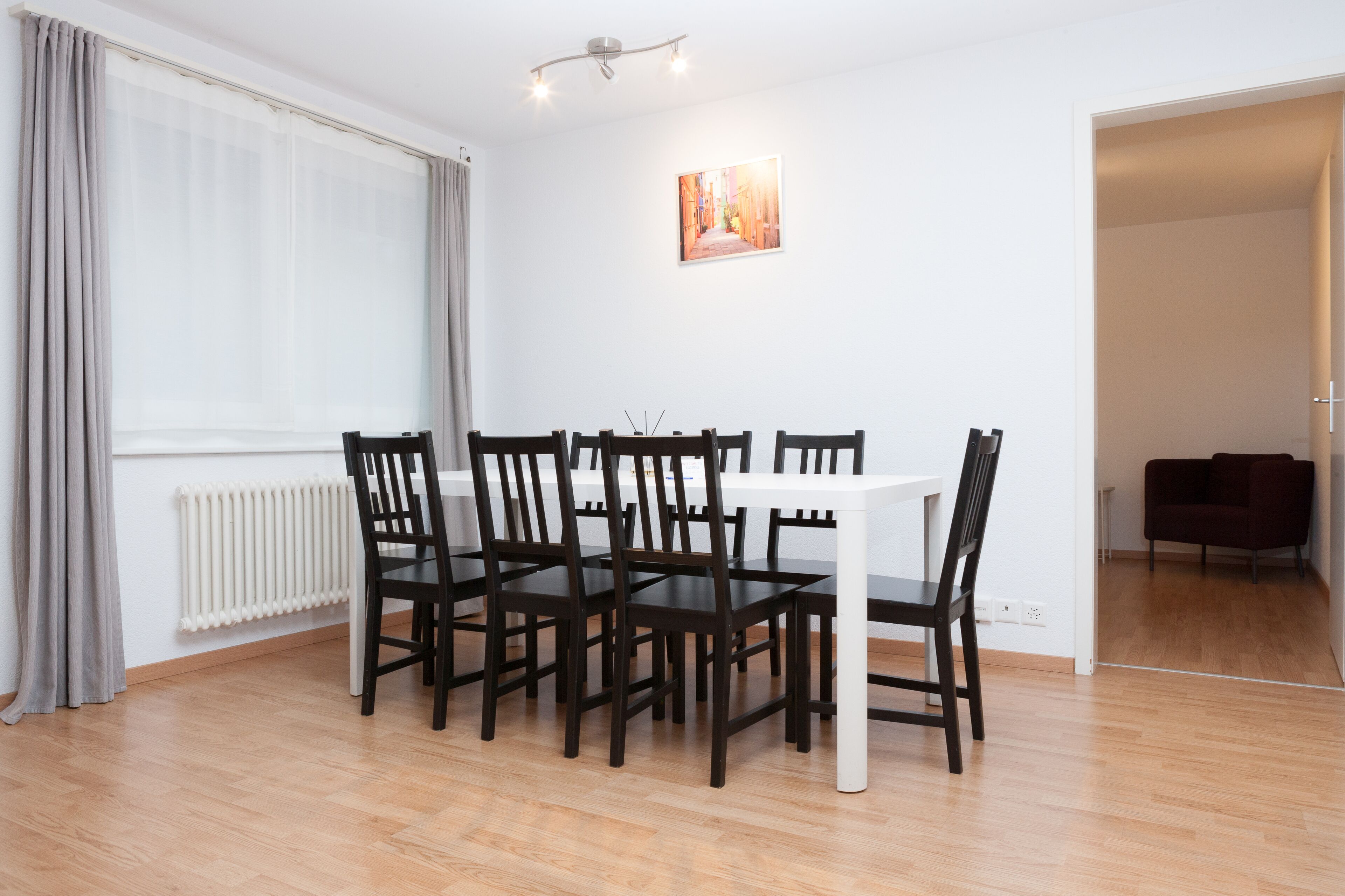 Ein Essbereich mit einem weißen Tisch und dunklen Holzstühlen, einem Heizkörper, einem Fenster mit Vorhängen und einem Gemälde an der Wand.