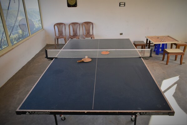 A ping pong table with a net, chairs, and a foosball table in a room with a large window. - Beyond Stay Tikker Narkanda Cottage (Theog)
