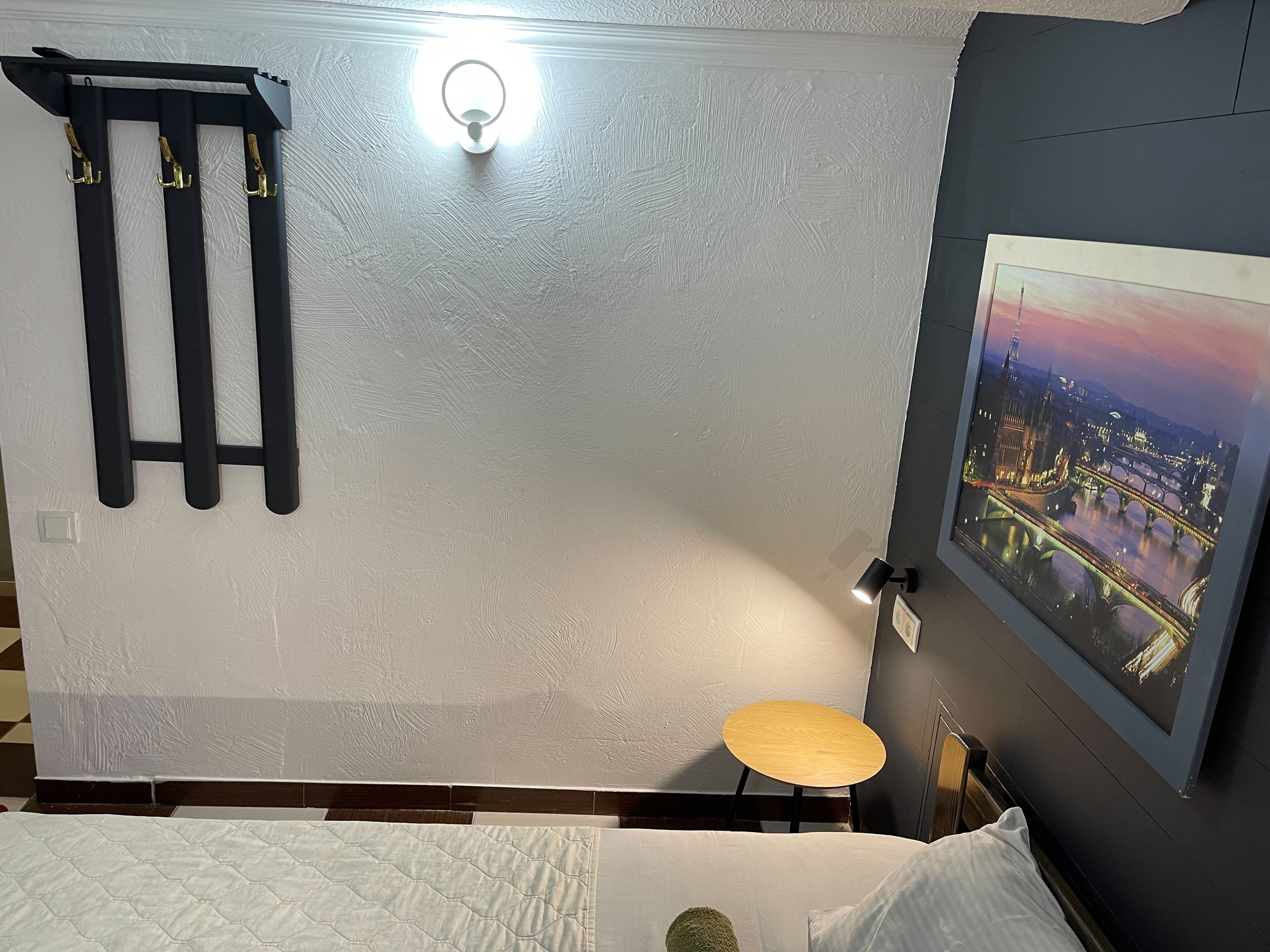 Une chambre d’hôtel avec un lit, une petite table ronde, une lampe fixée au mur et un tableau représentant une vue panoramique de la ville.
