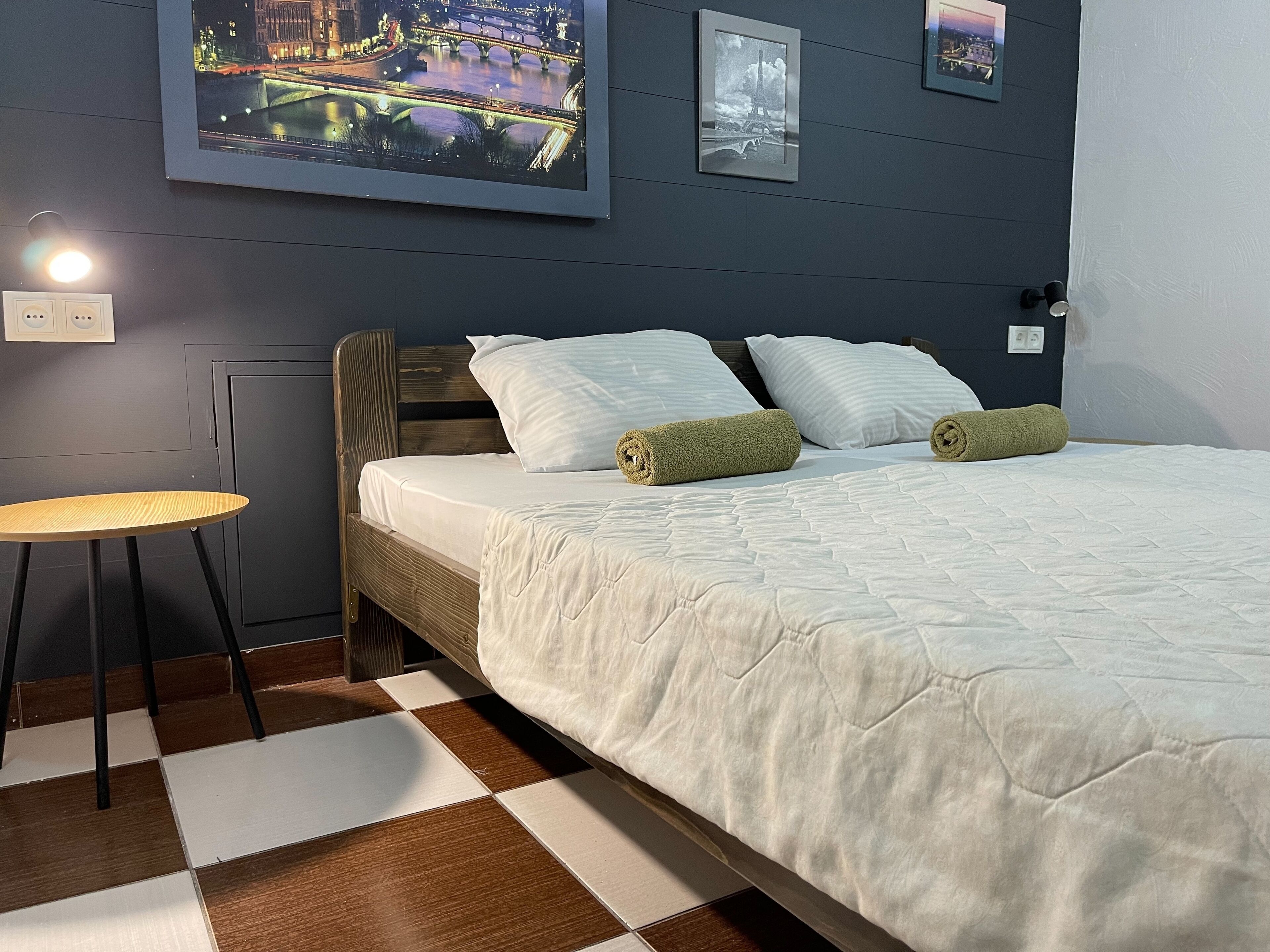 A neatly made bed with white bedding and two rolled towels, a small round table with a light, and a checkered floor.