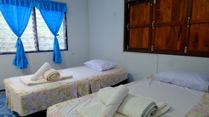 Une chambre avec deux lits, chacun agrémenté de draps et de serviettes à motifs floraux. Une fenêtre est ornée de rideaux bleus, et on peut apercevoir une porte en bois.