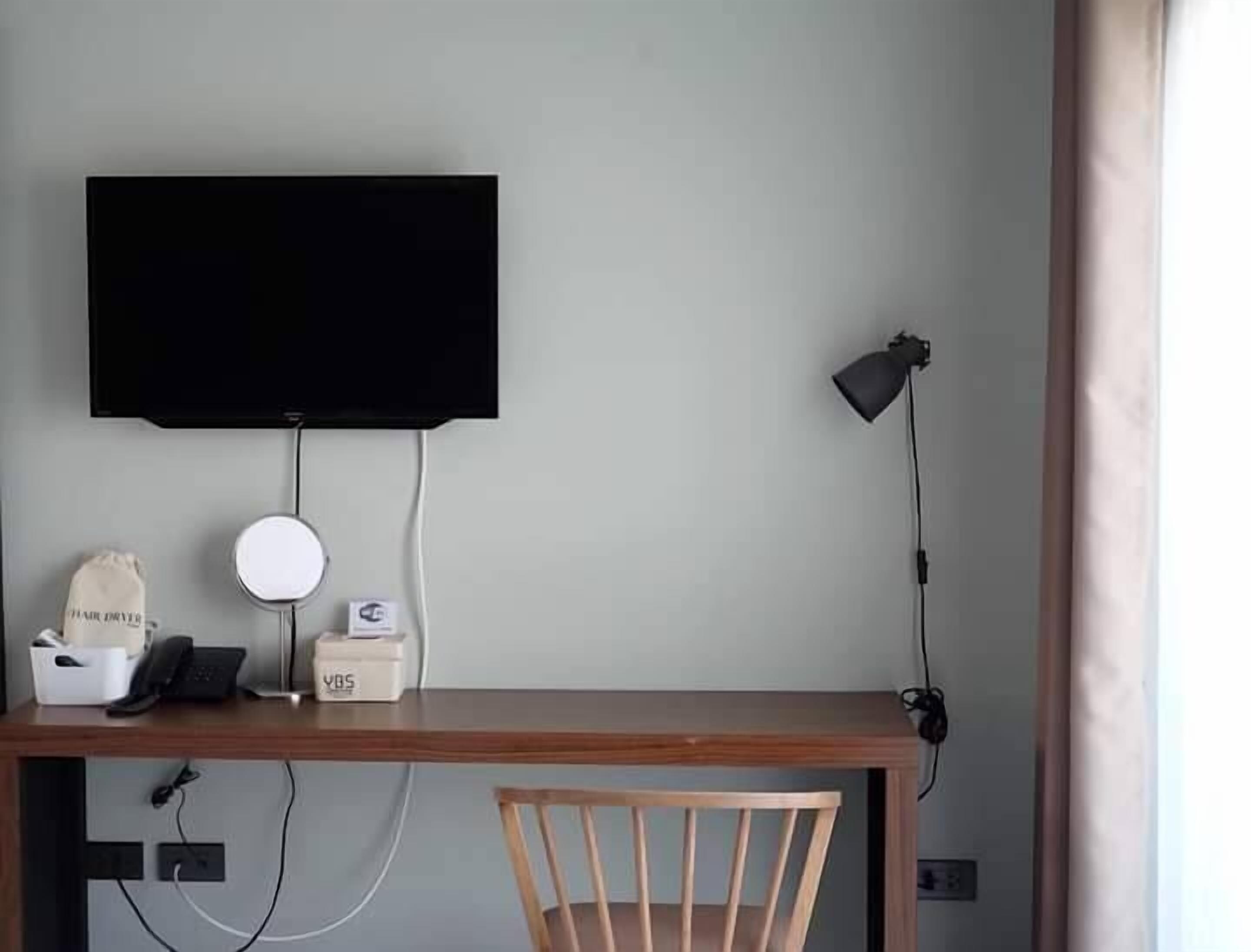 En arbetsplats med ett skrivbord i trä, en stol, en väggmonterad TV, en skrivbordslampa, en telefon, en rund spegel och en liten låda märkt med texten ”YBS”.