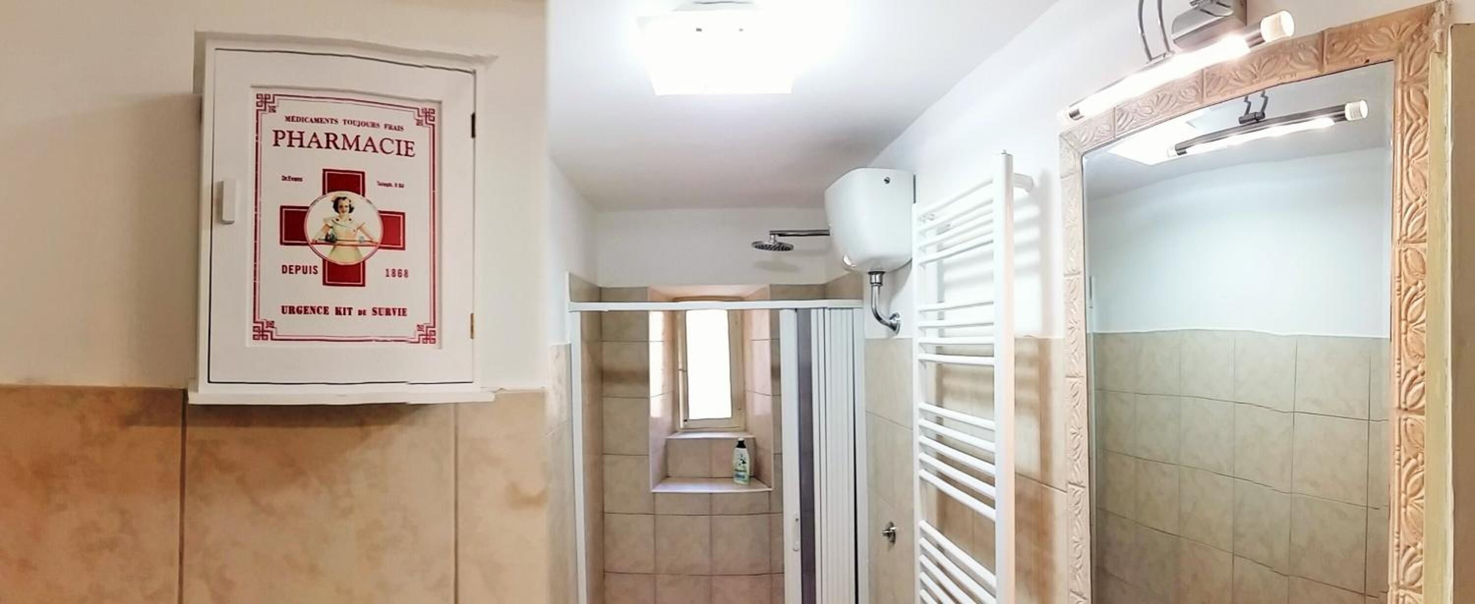 Baño con un mueble blanco que exhibe un cartel antiguo de farmacia, zona de ducha y espejo con un marco ornamentado.