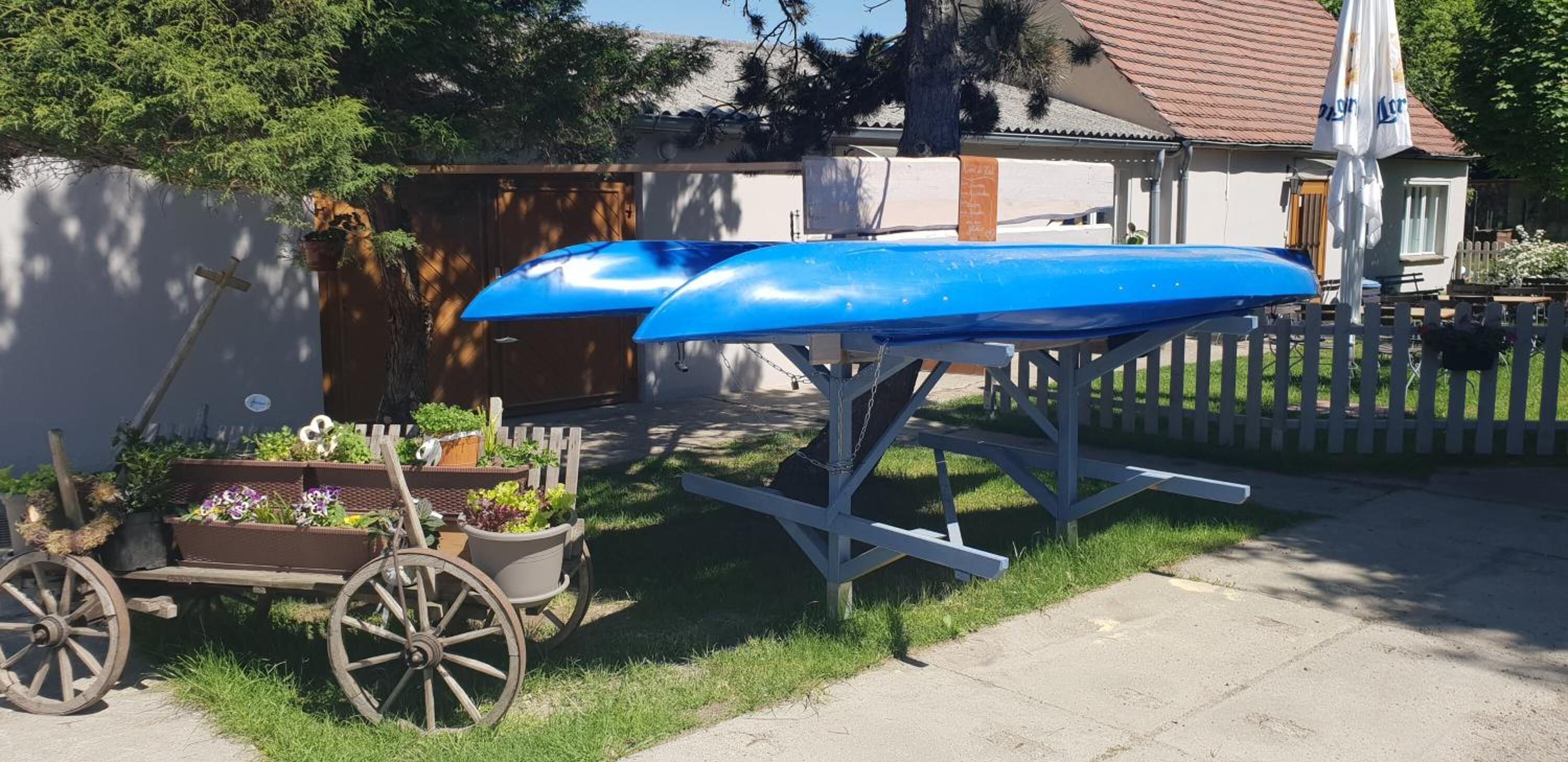 Un jardín con un carro de madera lleno de flores, una canoa azul sobre un soporte y una casa al fondo.