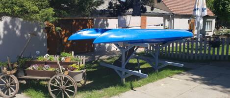 Un jardín con un carro de madera lleno de flores, una canoa azul sobre un soporte y una casa al fondo.