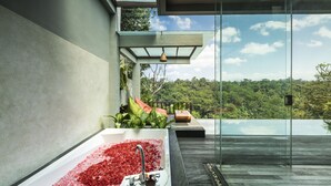 A modern bathroom with a large bathtub filled with rose petals, a glass-enclosed infinity pool, and a view of lush greenery. - Ayuterra Resort (Ubud)