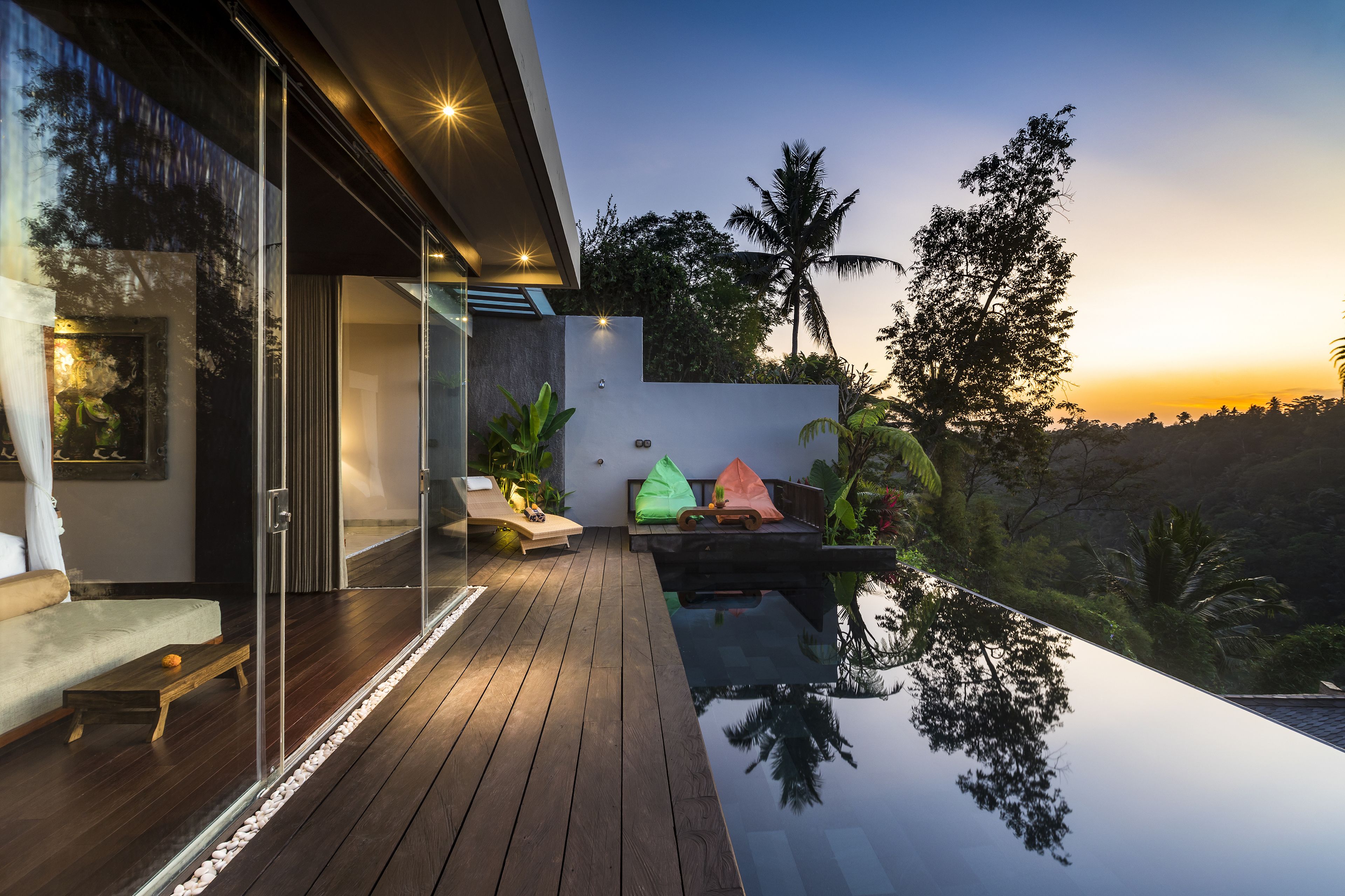 Una terraza moderna al aire libre con cerramiento de vidrio, piso de madera y una piscina que refleja los árboles circundantes.